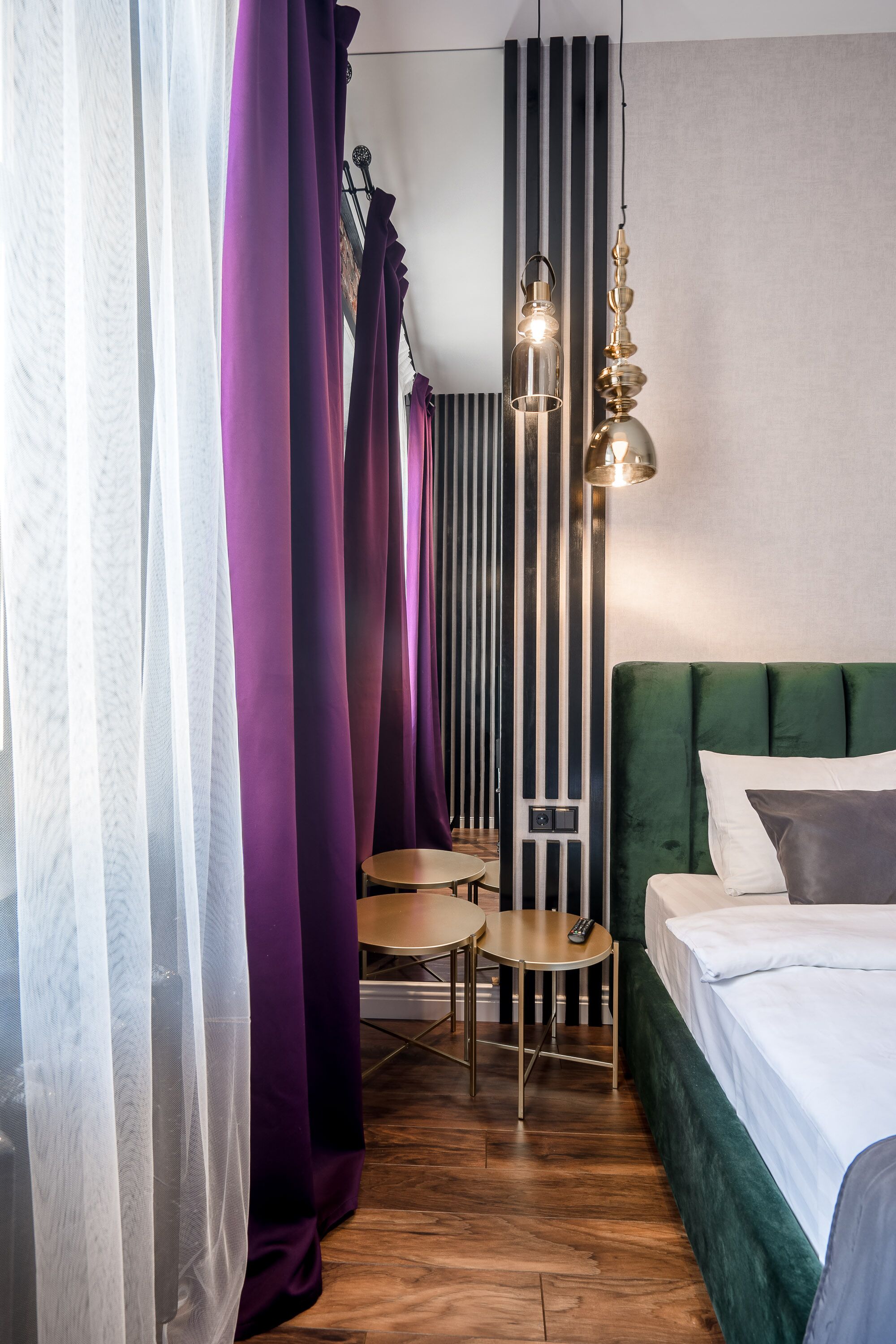 A hotel room with a green upholstered bed, a small round table, a chair, and purple curtains.