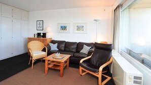 A living room with a black leather sofa, a wicker chair, a wooden coffee table, and a white radiator. There are three framed pictures on the wall and a window with blinds. - Apartment 91 Against the wind (ID 012) (Sankt Peter-Ording)