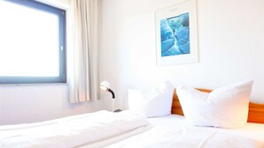 A hotel room with a bed, a bedside lamp, a window with curtains, and a framed picture on the wall. - Apartment 91 Against the wind (ID 012) (Sankt Peter-Ording)