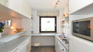 A compact kitchen with white cabinets, a microwave, a sink, and a window with a curtain. - Apartment 91 Against the wind (ID 012) (Sankt Peter-Ording)