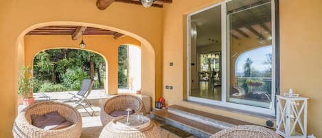 Un área de estar al aire libre cubierta, con mobiliario de mimbre y un amplio ventanal con vista a un jardín.