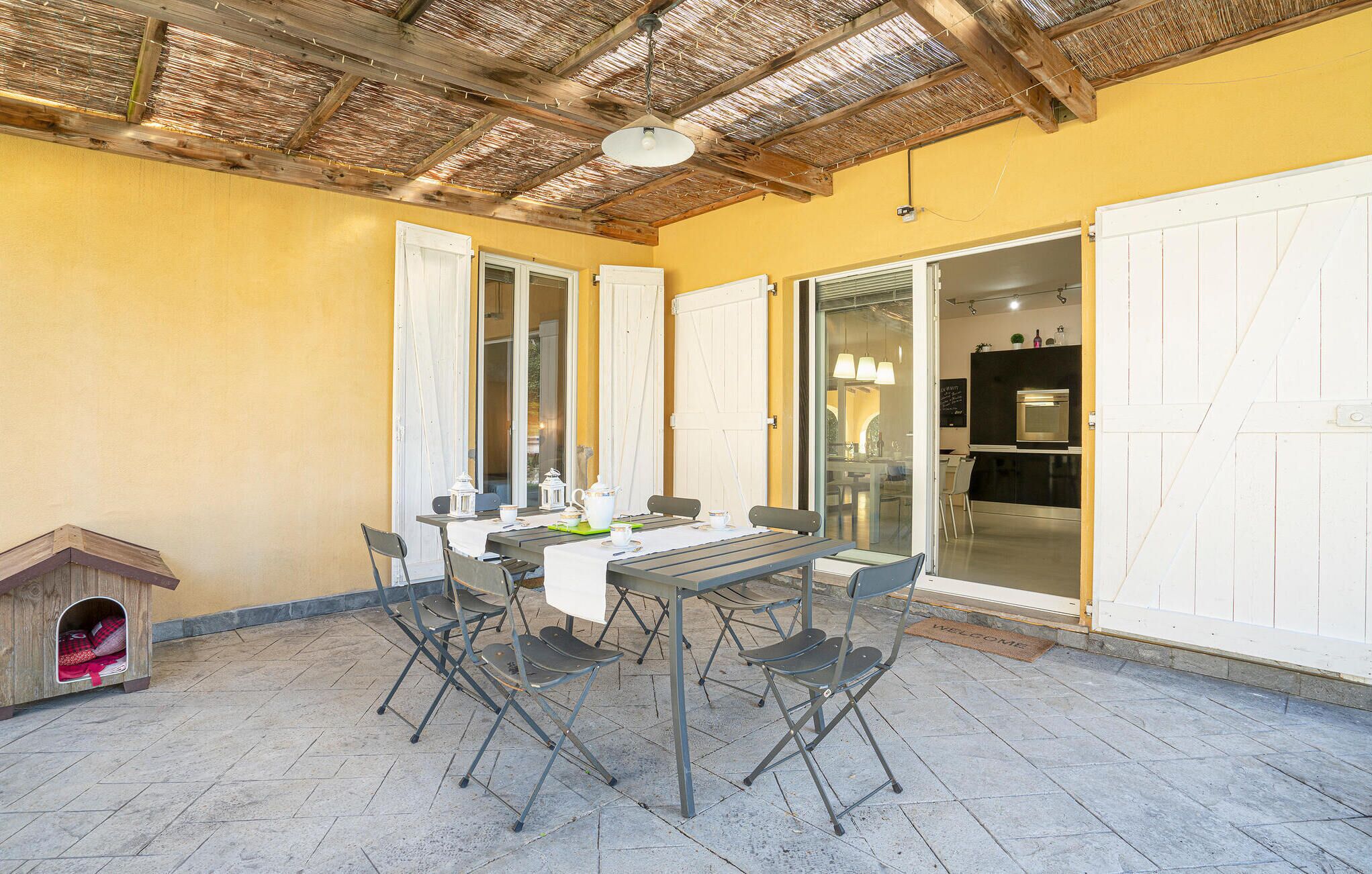 Une terrasse avec une table et des chaises, une niche pour chien et une cuisine visible à travers une porte ouverte.