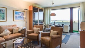 A hotel room with a sofa, armchairs, a glass coffee table, a dining area with a table and chairs, and a large window with a view. - Apartment 187 (ID 093) (Sankt Peter-Ording)