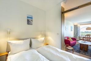 Quarto de hotel com cama, abajures, uma mesinha e vista para uma área de estar externa.
