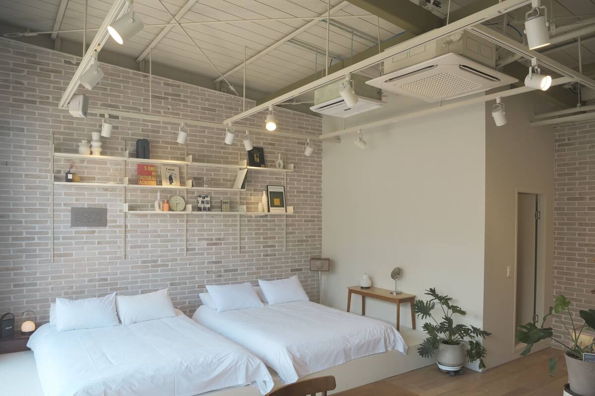 Moderni hotellihuone, jossa on kaksi sänkyä, puinen pöytä ja tiiliseinä, jossa on hyllyjä.