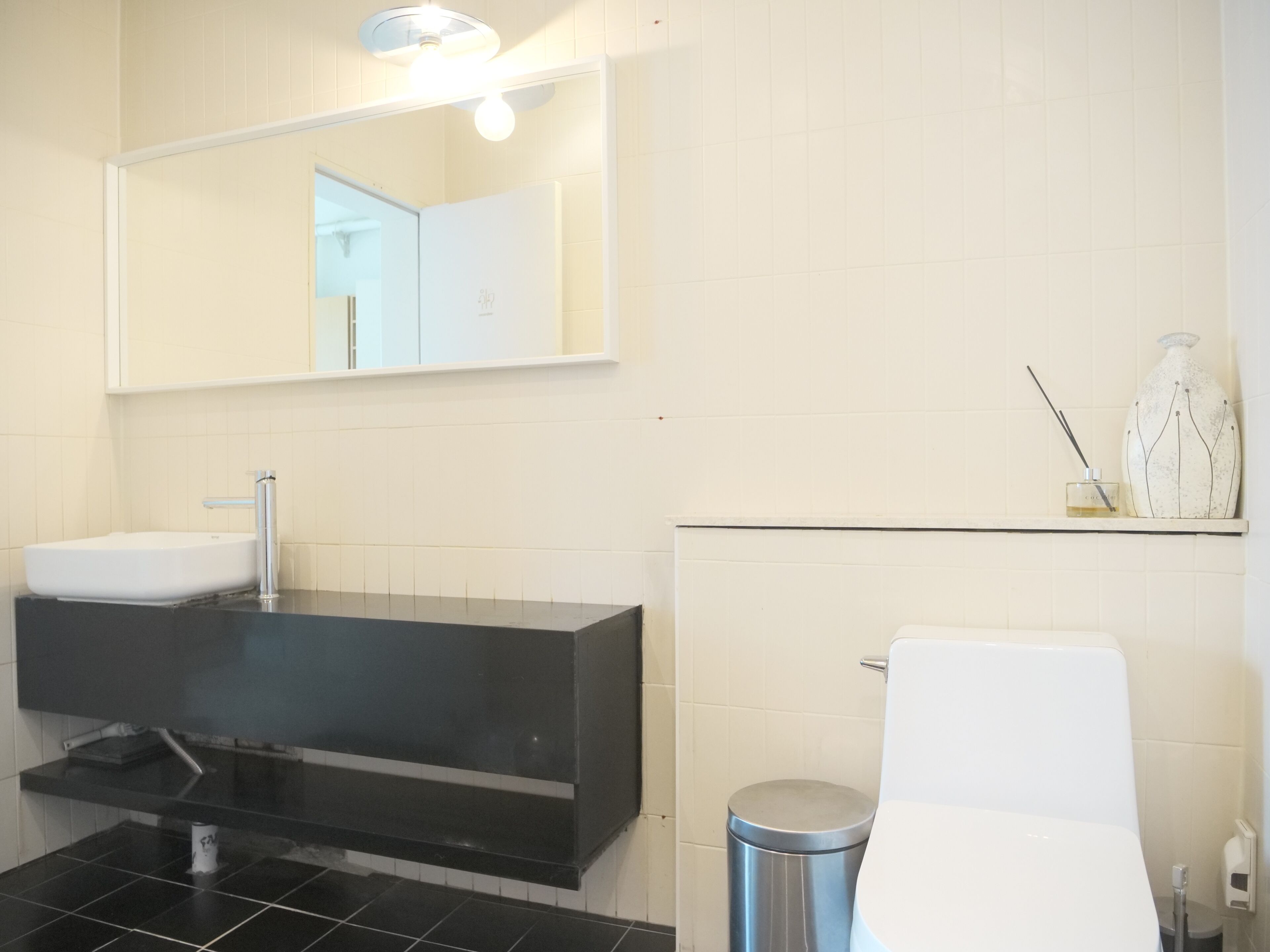 A modern bathroom with a white sink, black countertop, and a mirror above. There is a toilet with a white seat and a trash can beside it.