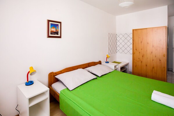A bedroom with a green bedspread, wooden headboard, white pillows, a framed picture on the wall, a wooden wardrobe, and a small white nightstand with a lamp.