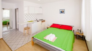 A bedroom with a green bedspread, a ceiling fan, a small kitchenette, and a view of a green outdoor area. - Friendly People's Guest House (Zadar)