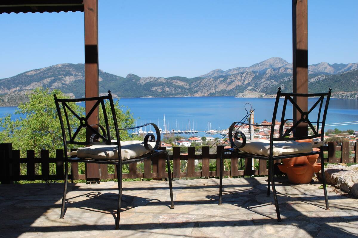 Deux chaises en métal avec des coussins, placées sur une terrasse en pierre donnant sur une étendue d’eau où se trouvent des bateaux, avec un paysage montagneux en arrière-plan.