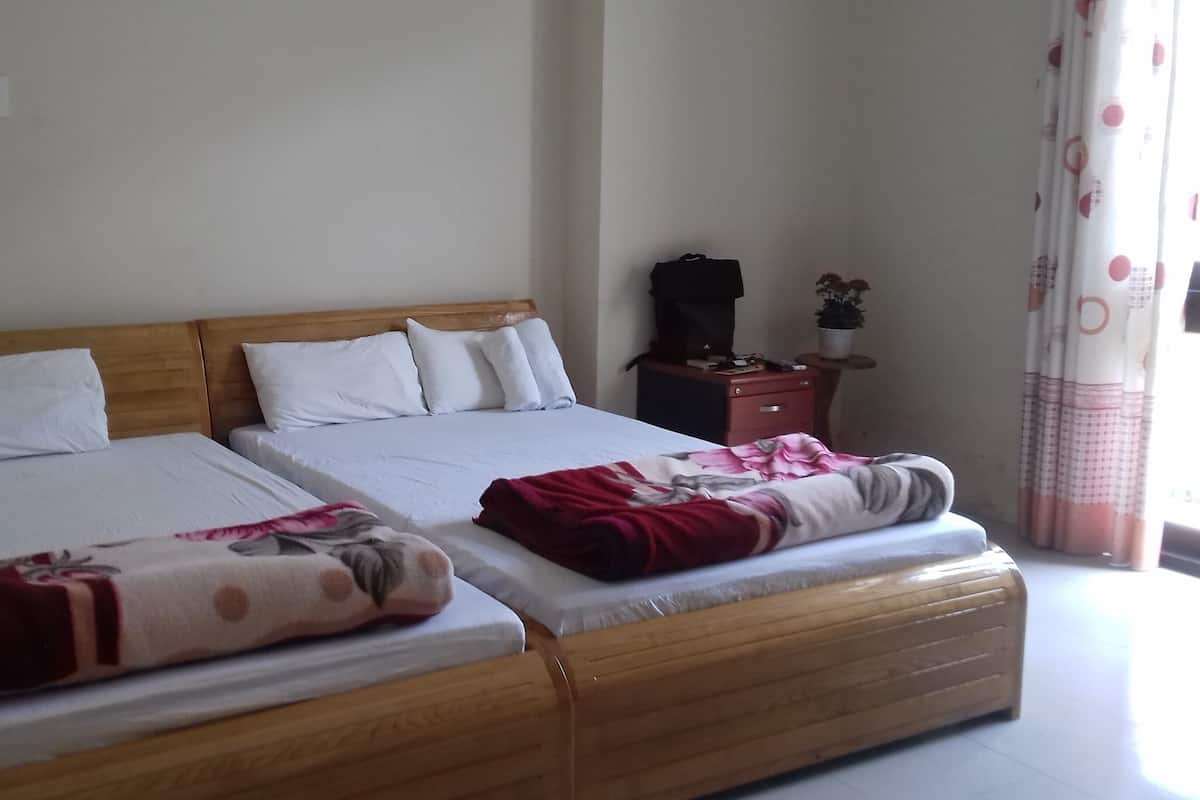 A double bed with a wooden headboard, two white pillows, and a red and white blanket. There is a small wooden nightstand with a plant and a black bag on it.