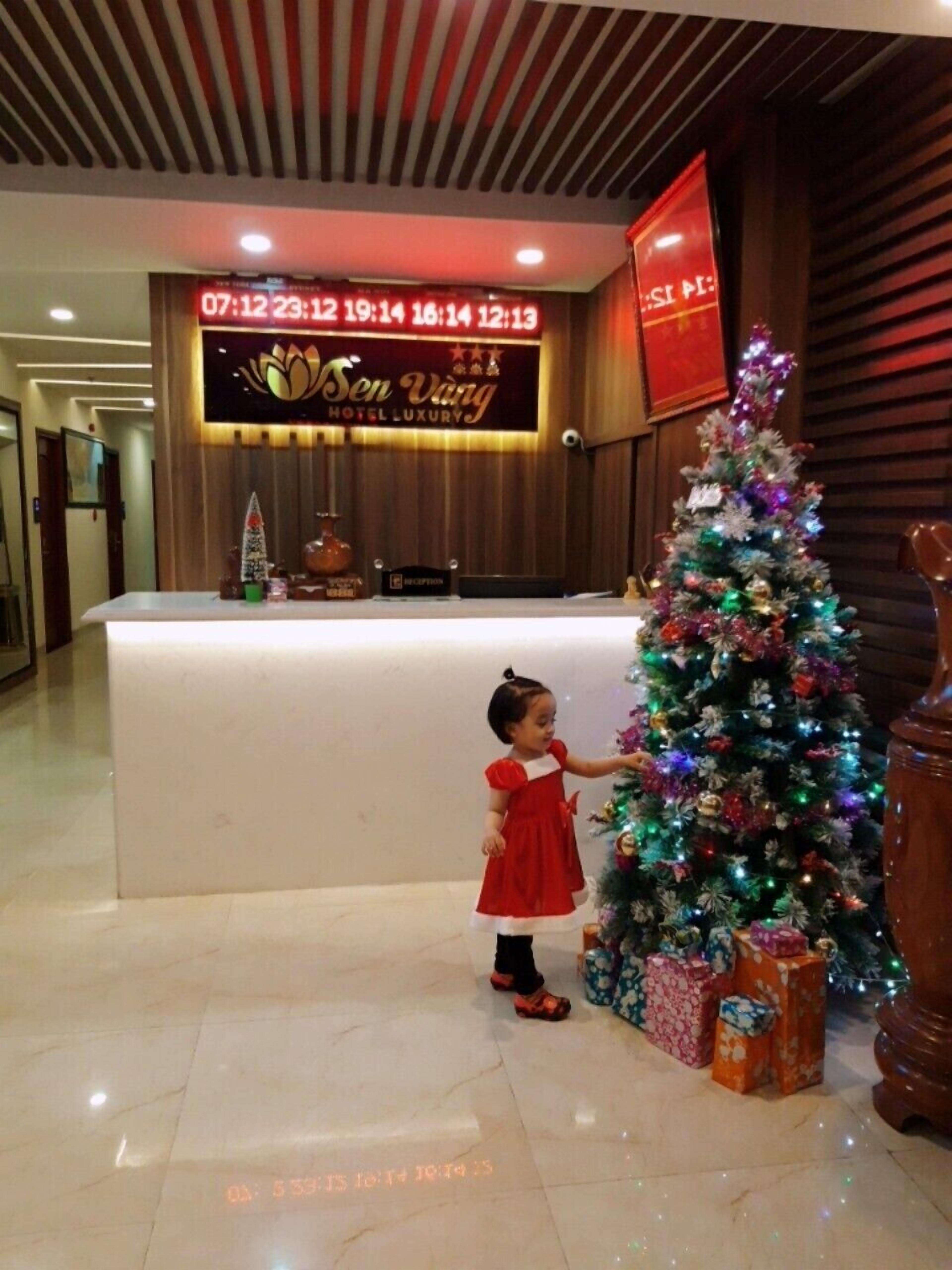 A reception desk with a digital clock displaying hotel hours, a Christmas tree with gifts, and a child in a red dress.