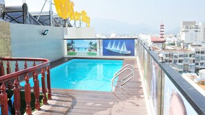 A rooftop pool with a wooden deck and metal railings, surrounded by city buildings and a clear sky.