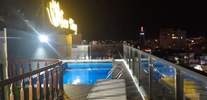 Zona de piscina en la azotea con barandilla de vidrio, un letrero de neón y vista panorámica de la ciudad al fondo.