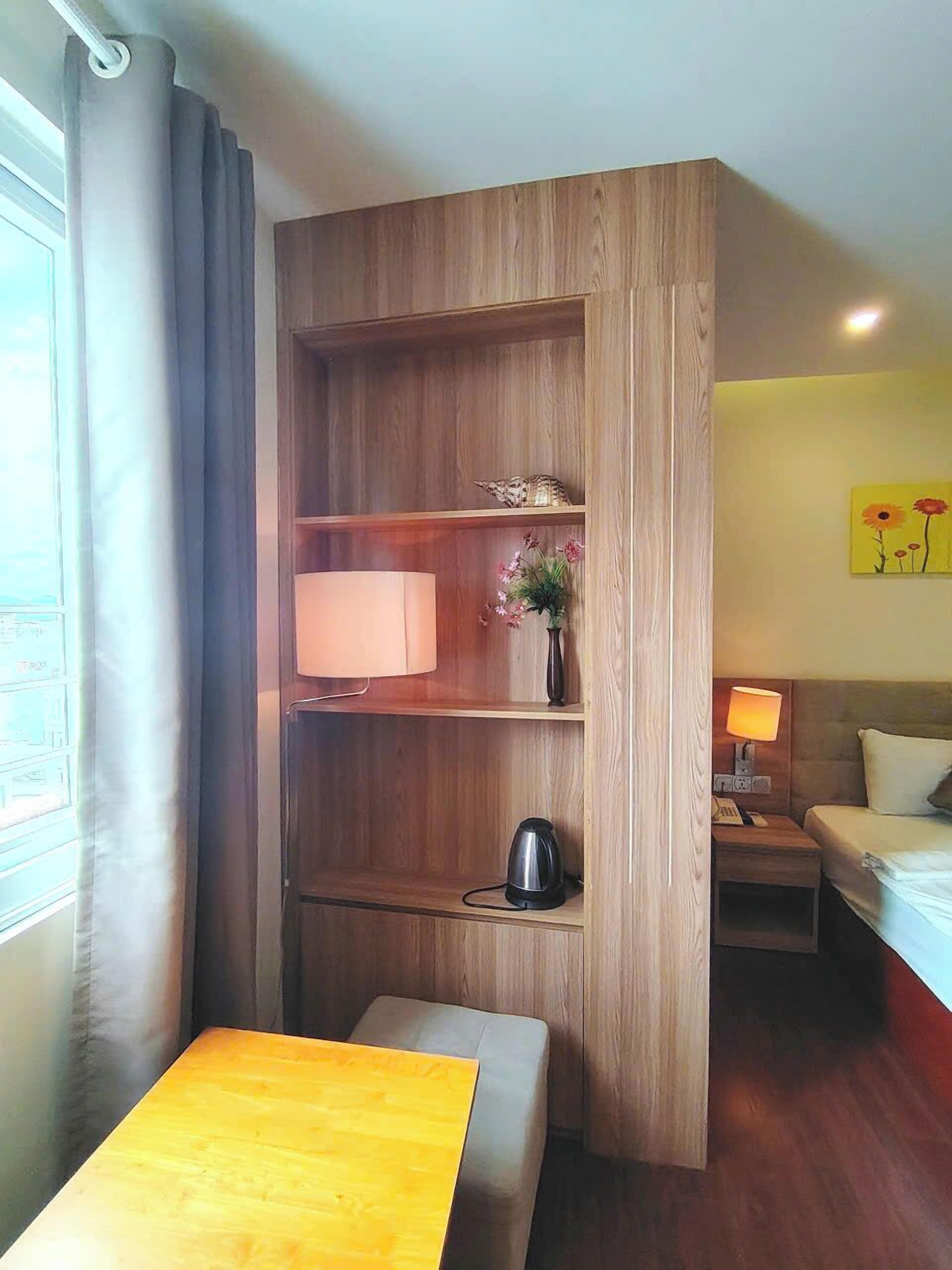 A hotel room with a wooden cabinet, a bedside table, a bed, a small table with a lamp, and a painting on the wall.