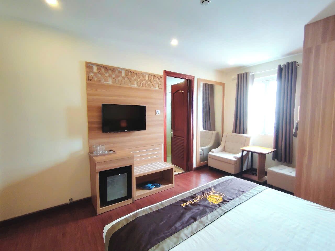 Een hotelkamer met een bed, een aan de muur bevestigde televisie, een zithoek met een stoel en een tafeltje, en een raam met gordijnen.