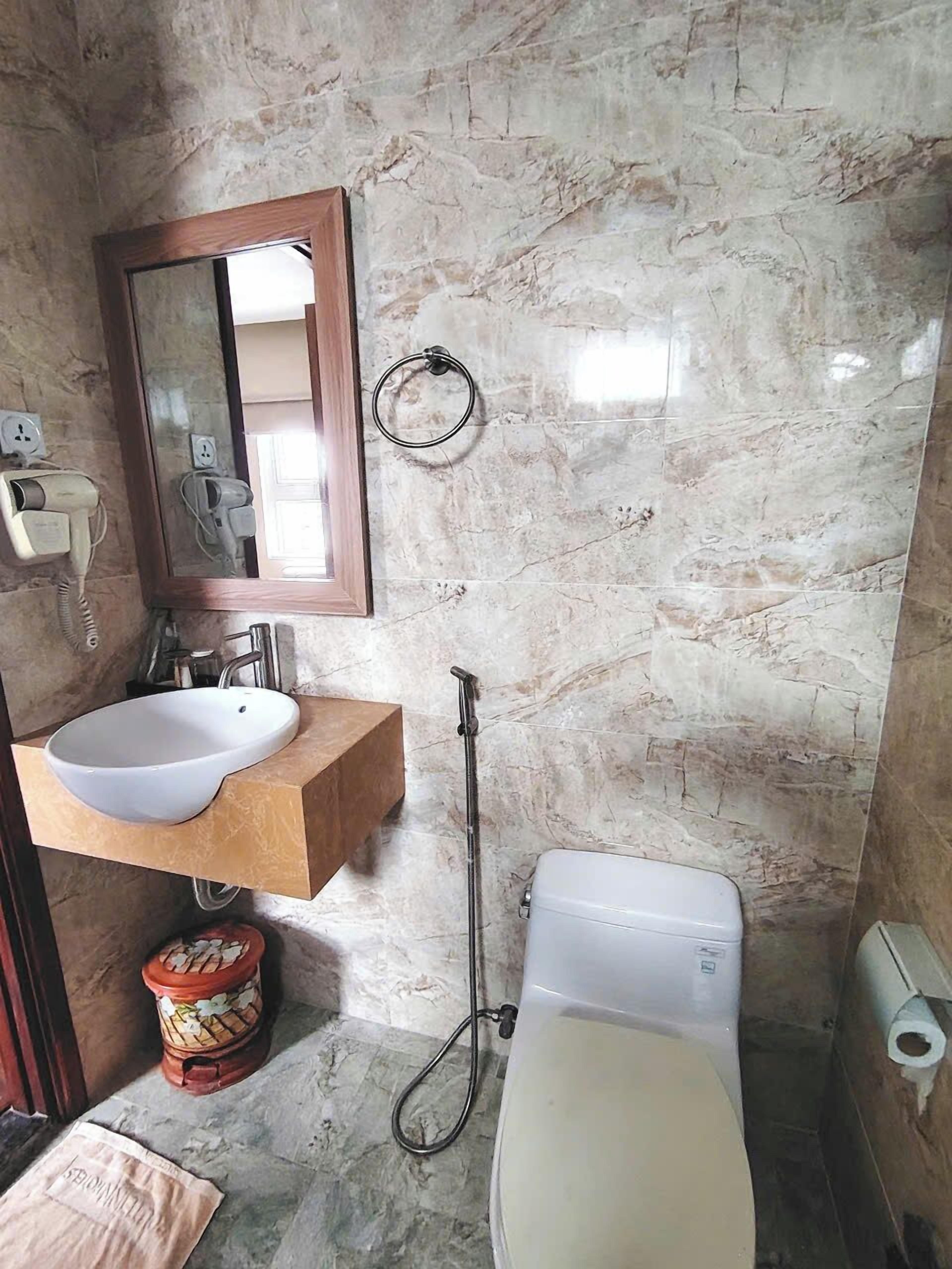 A bathroom with a marble wall, a wooden vanity with a white sink, a mirror, a toilet, and a shower hose.