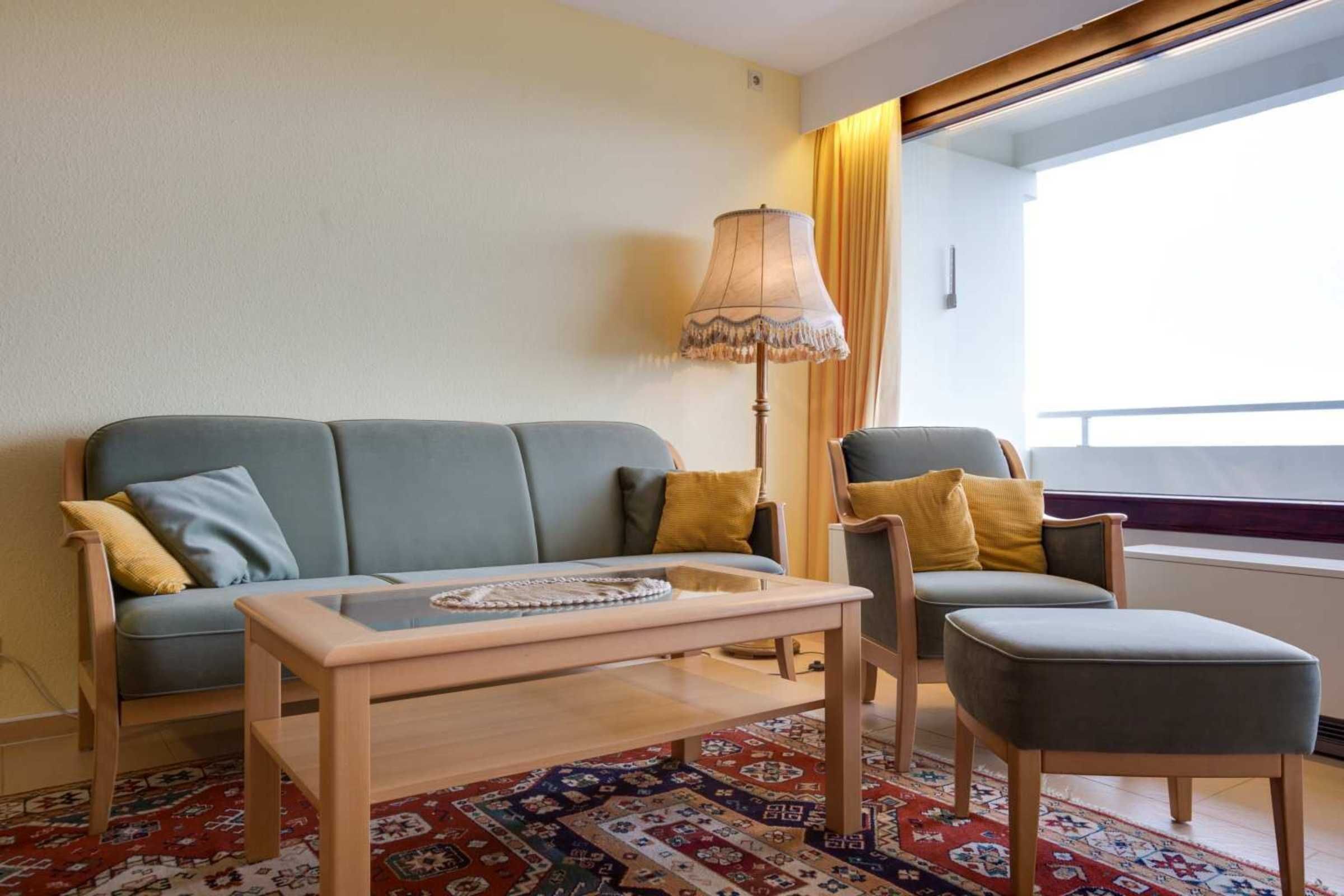 A living room with a sofa, armchair, ottoman, and a table with a lamp.