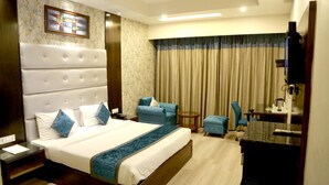 A hotel room with a large bed, a desk, a chair, a small table, a television, and a ceiling fan. - Hotel Shagun (Dera Bassi)