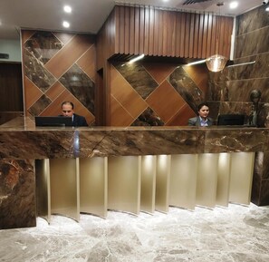 Una recepción con paneles de mármol y madera, dos empleados y una estatua.