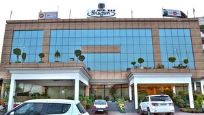 A modern hotel with a large glass facade, a prominent sign reading "Hotel Shagun," and a parking area with several cars. - Hotel Shagun (Dera Bassi)