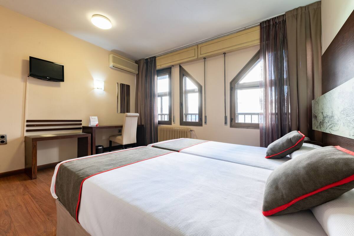 Habitación de hotel con dos camas, un escritorio, una silla, un televisor y ventanas grandes con cortinas.