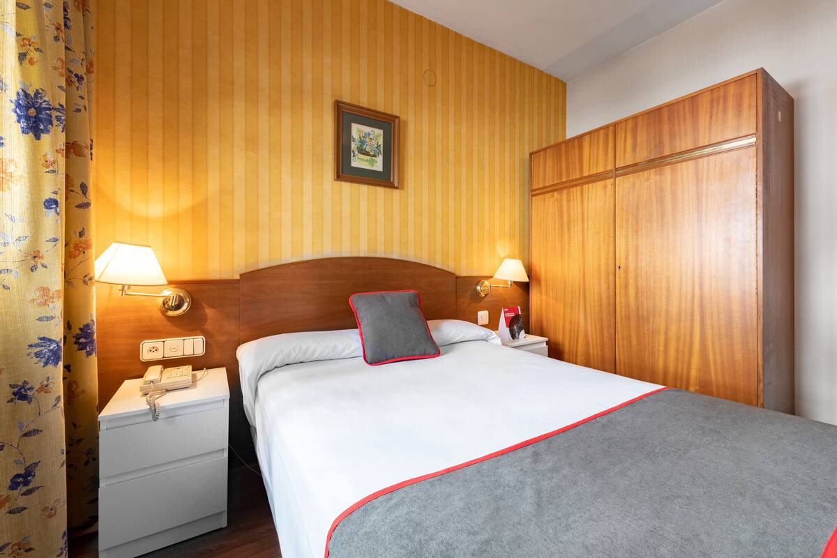 Habitación de hotel con cabecera de madera, cama con manta gris, dos lámparas de noche, mesita de noche con teléfono y armario de madera.
