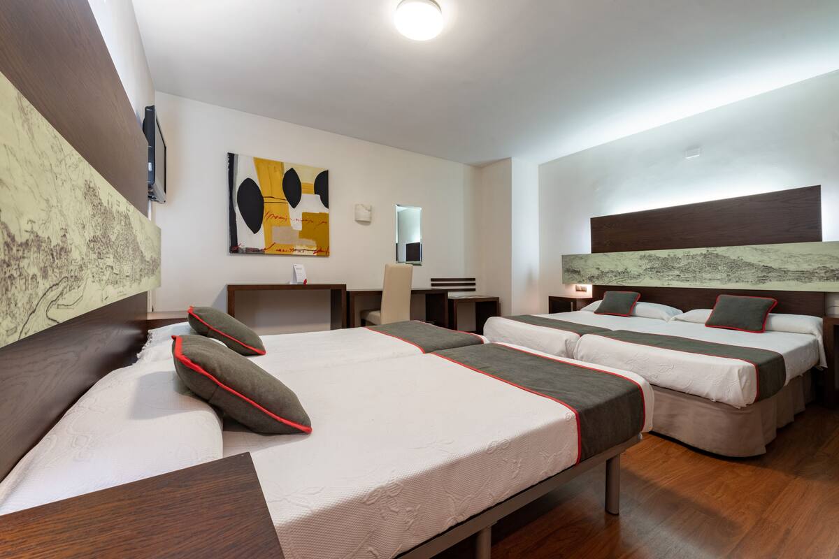 Habitación de hotel con tres camas, un escritorio, una silla y obras de arte en las paredes.