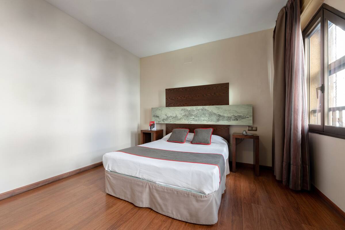 Habitación de hotel impecable con una cama grande, cabecera con panel decorativo, mesita de noche con lámpara y ventana con cortinas.