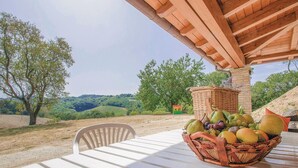 Une table surmontée d’un panier en osier rempli de fruits, placée sous une pergola en bois, d’où l’on peut admirer un paysage vallonné parsemé d’arbres.