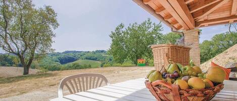 Een tafel met een rieten fruitmand, onder een houten pergola, met uitzicht op een glooiend landschap met bomen.