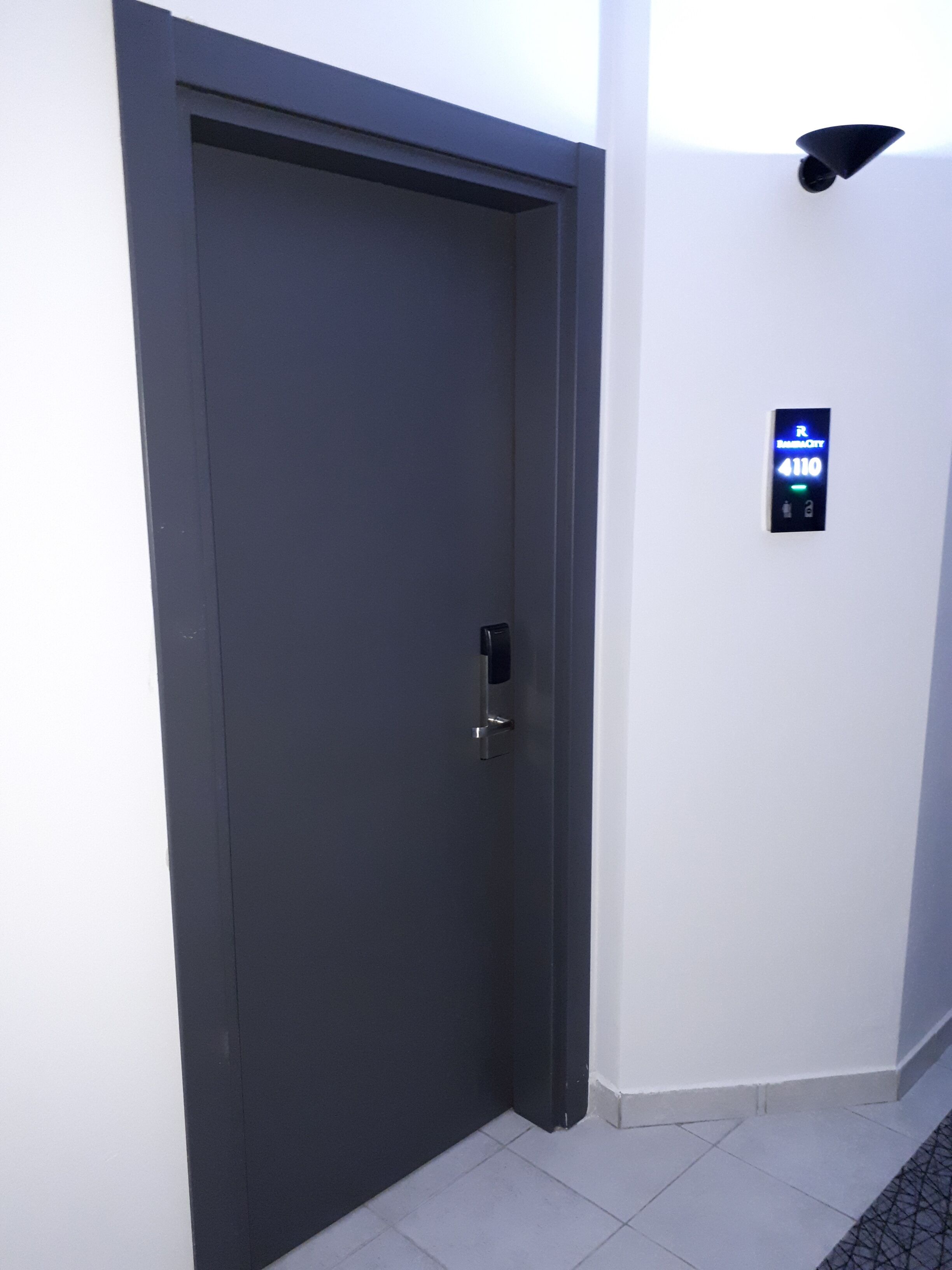 A dark grey door with a keypad lock, a small illuminated sign above it, and a tiled floor.