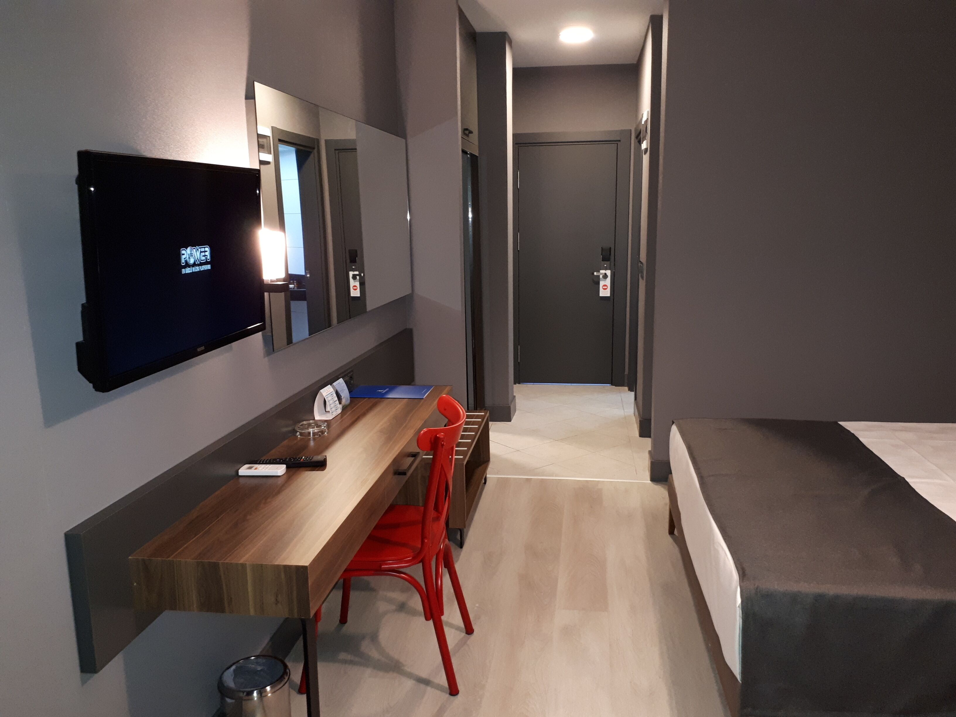 A modern hotel room with a wooden desk, red chairs, a flat-screen TV, a mirror, and a bed.