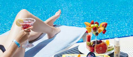 A poolside setting with a person relaxing on a lounge chair, holding a drink, and a fruit platter nearby.