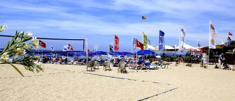 A beach volleyball court with flags, sun umbrellas, and beach chairs.