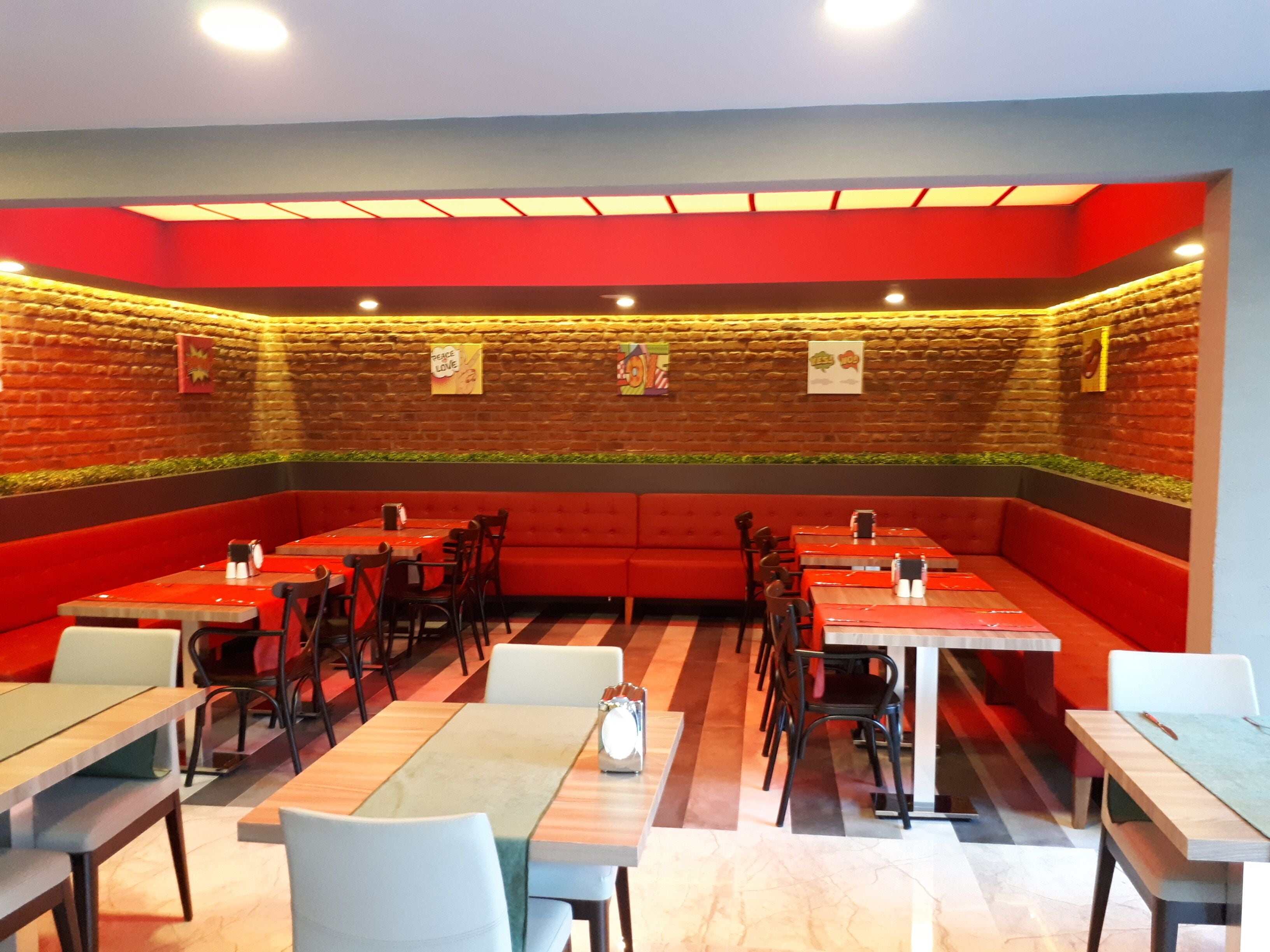 Un restaurante con asientos y mesas rojas, paredes de ladrillo y una franja de plantas verdes.