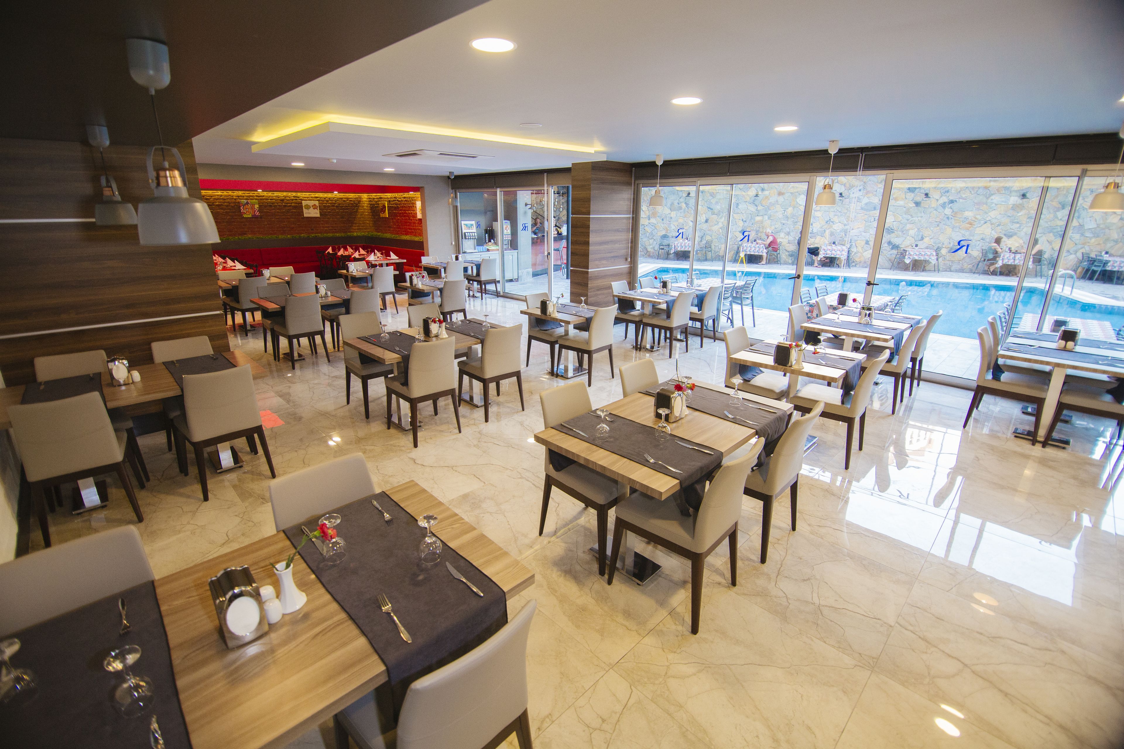A spacious restaurant with multiple dining tables and chairs, a pool area visible through large windows, and a modern interior design.