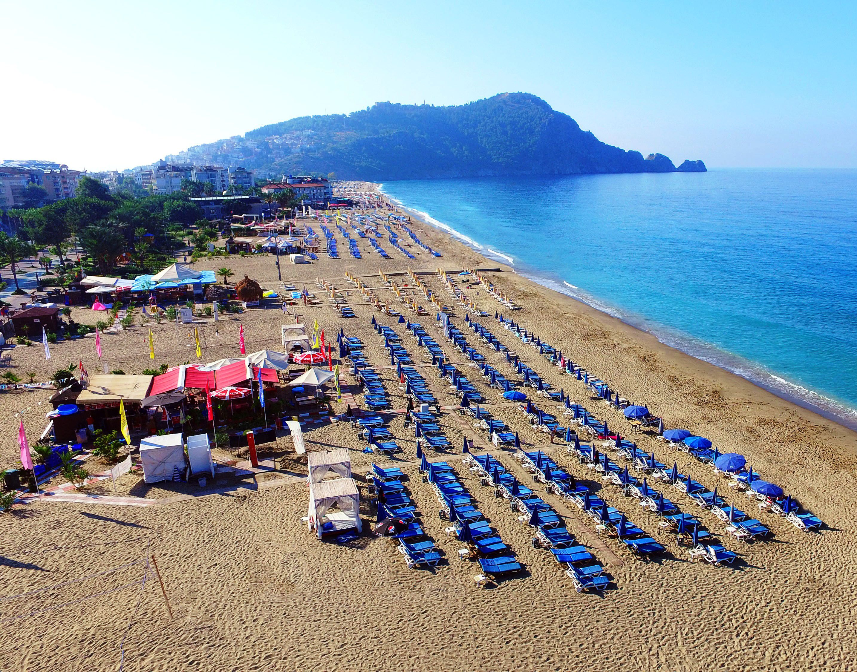 A beach with rows of blue umbrellas and sun loungers, a few beachside shops, and a hilly landscape in the background.