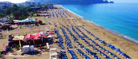 A beach with rows of blue umbrellas and sun loungers, a few beachside shops, and a hilly landscape in the background.