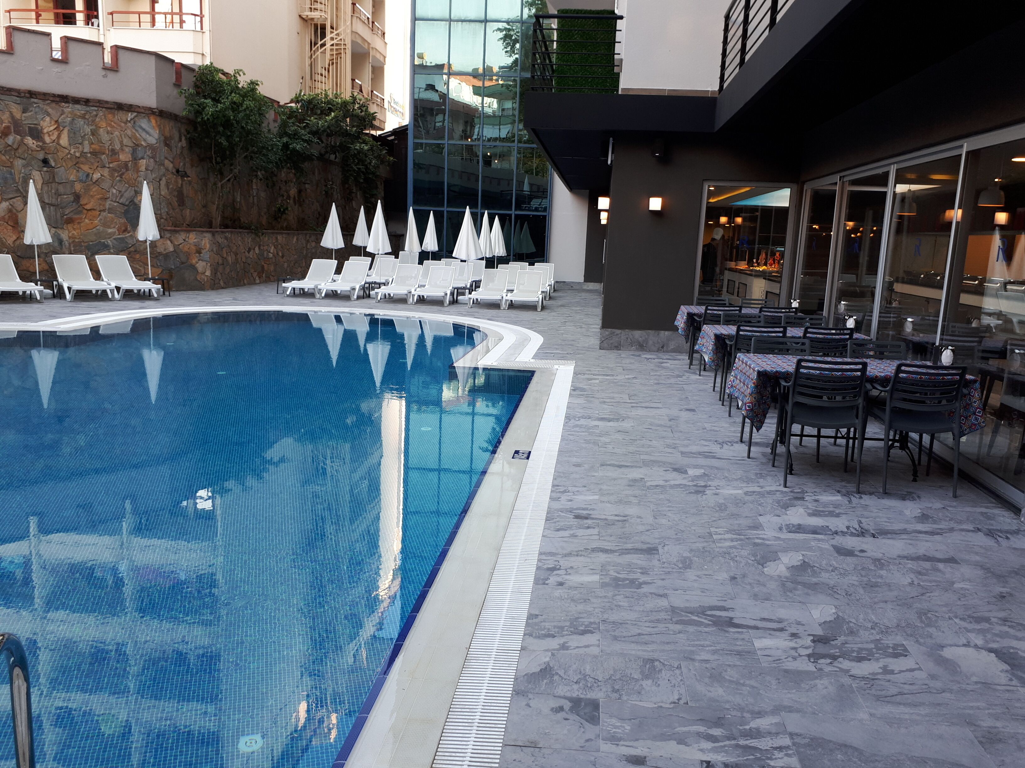 A swimming pool with lounge chairs and tables, surrounded by stone walls and a glass building.