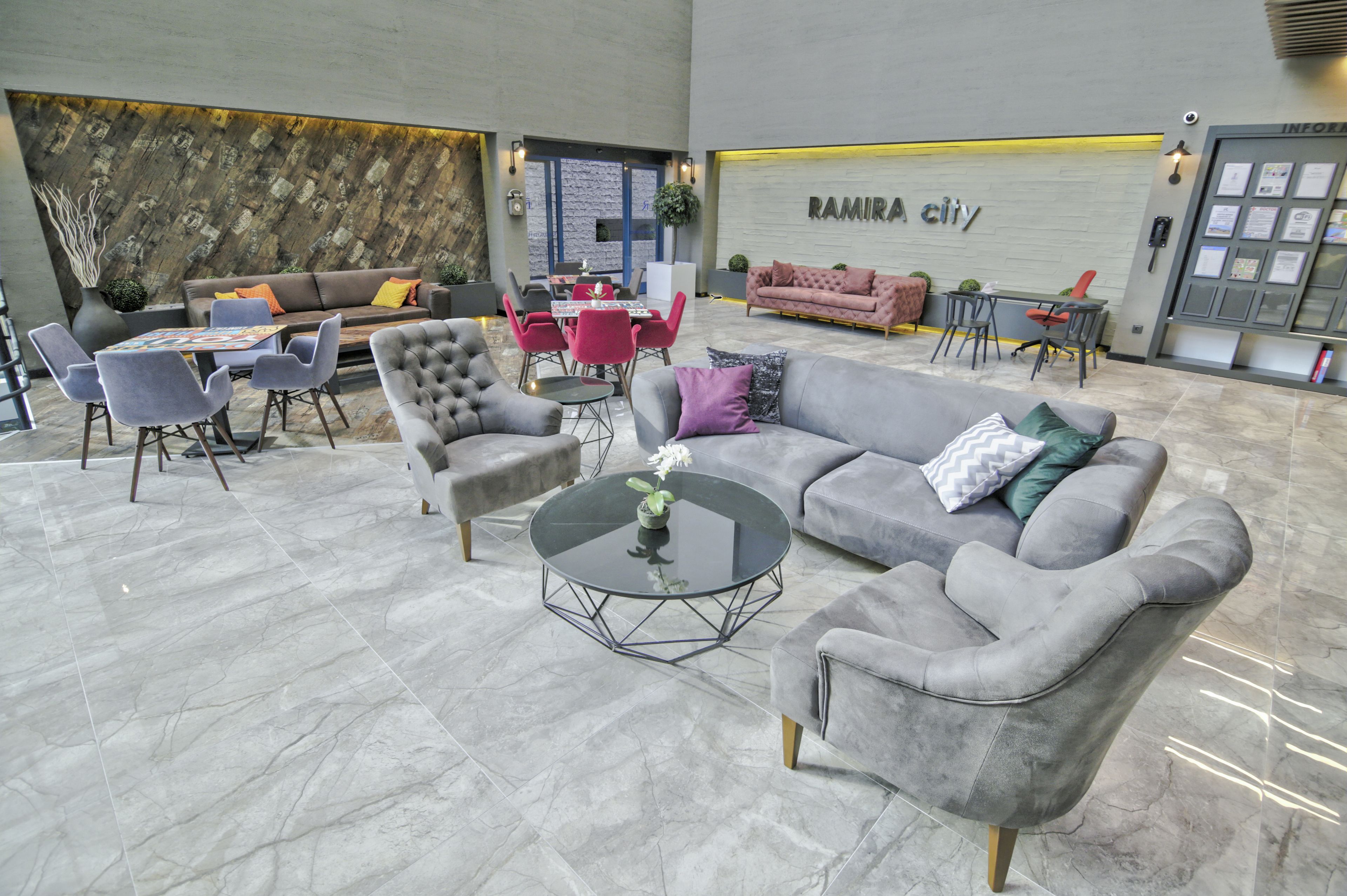 Un vestíbulo amplio con un sofá gris, sillones y una mesa de centro. Se ve una recepción con una computadora y un área de espera con sillas y una mesa.