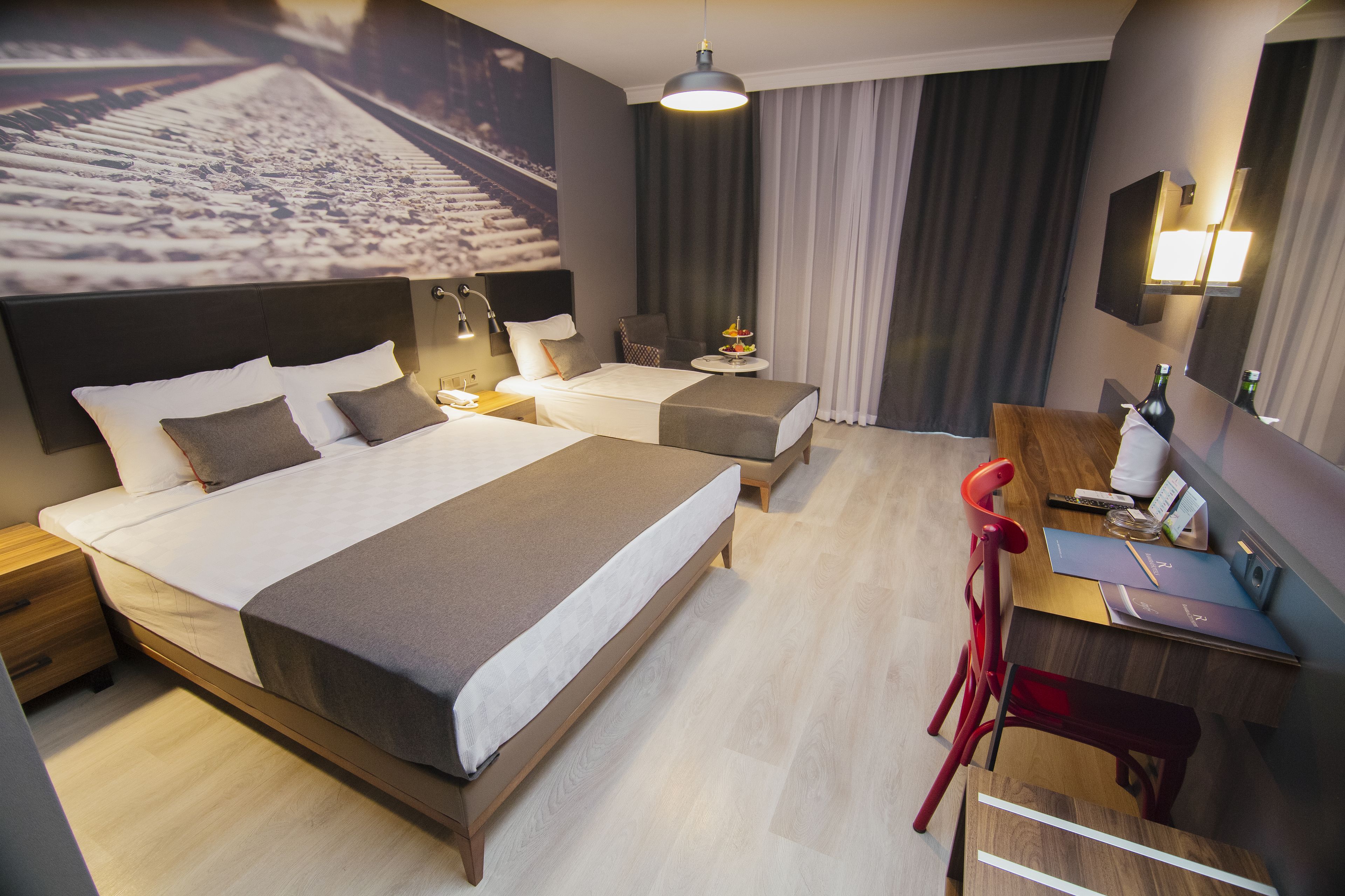 Ein Hotelzimmer mit einem großen Bett, einem kleineren Bett, einem Schreibtisch mit Stuhl, einem Nachttisch und einem an der Wand befestigten Fernseher.