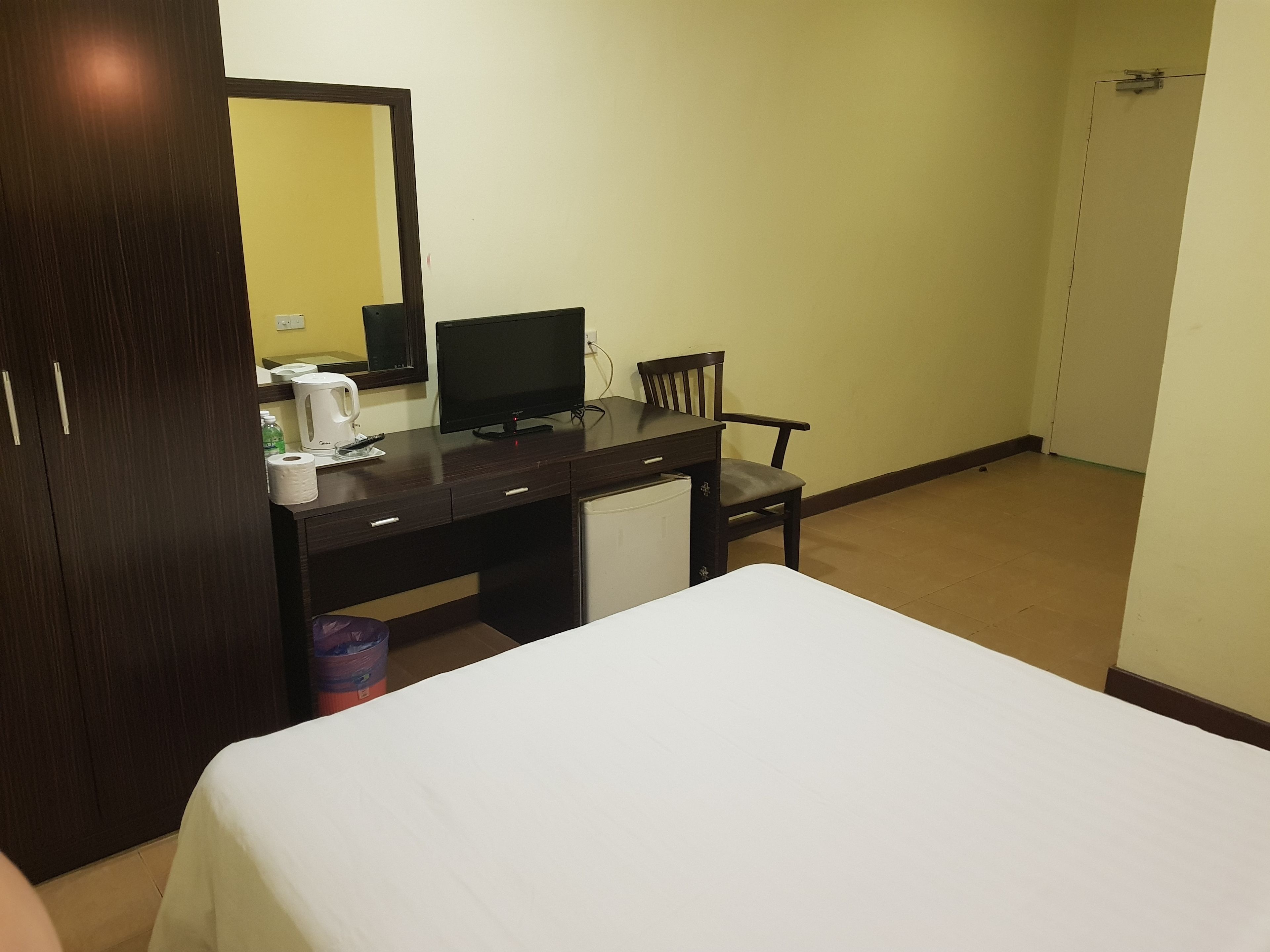 A hotel room with a bed, a desk with a computer, a chair, a small refrigerator, and a mirror.