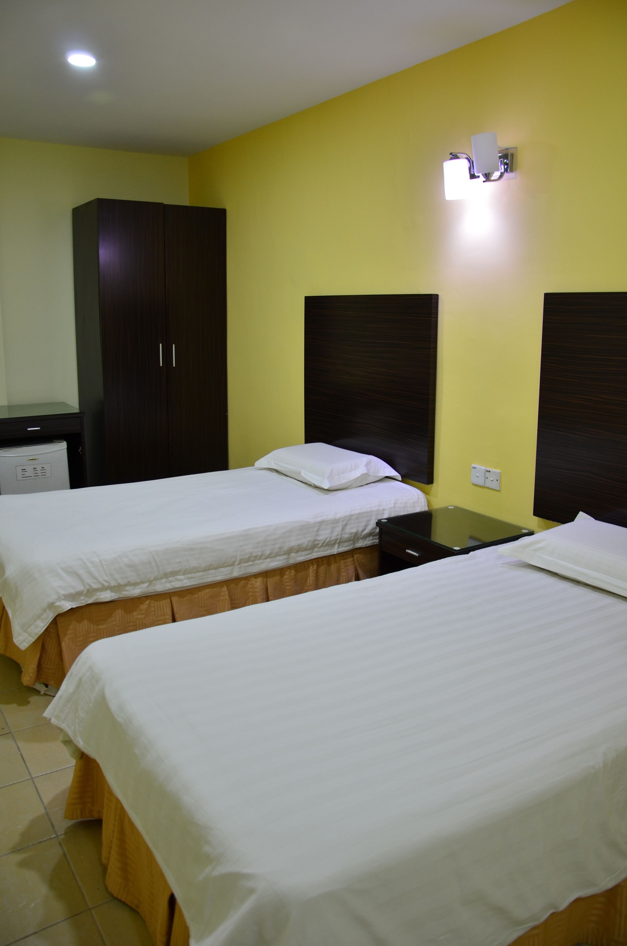 A hotel room with two beds, a wardrobe, a desk, and a wall-mounted light fixture.