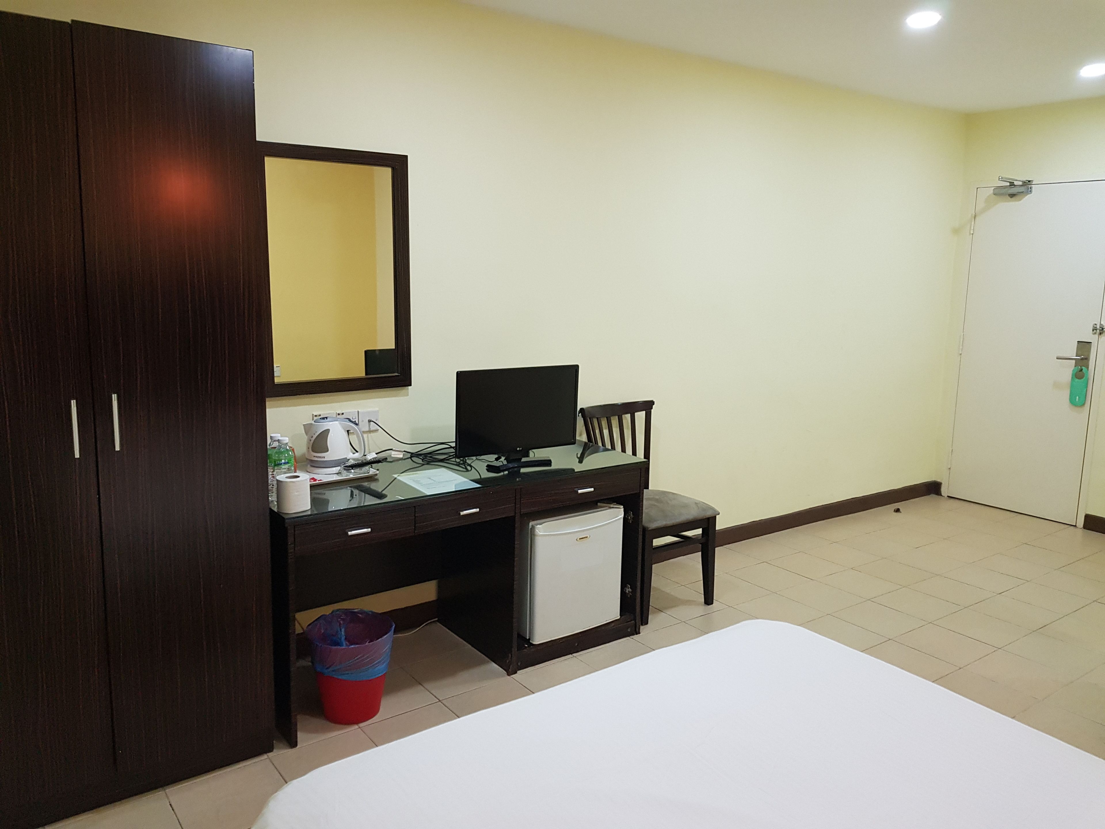 A hotel room with a bed, a desk with a computer, a chair, a small refrigerator, a trash bin, and a wardrobe.