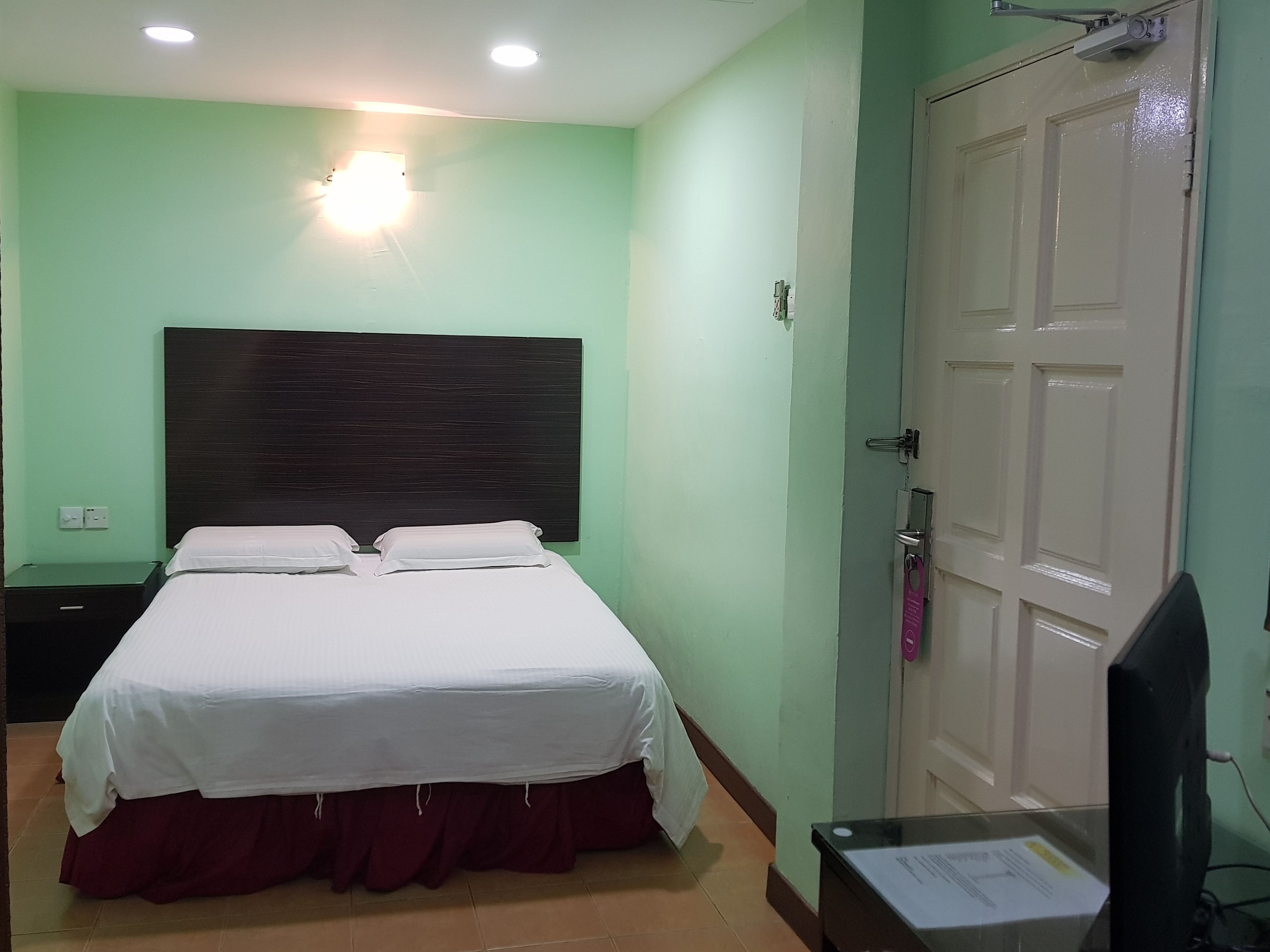 Quarto de hotel com uma cama, mesa de cabeceira, escrivaninha e um computador, além de uma porta.