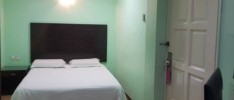 Quarto de hotel com uma cama, mesa de cabeceira, escrivaninha e um computador, além de uma porta.