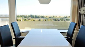 Une salle de conférence avec une longue table et quatre chaises, de grandes fenêtres donnant sur un paysage verdoyant et un plan d’eau, et une horloge au mur.