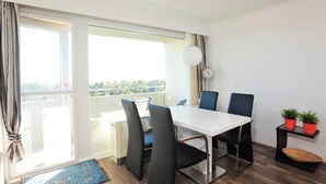 Un espace repas moderne avec une table blanche et des chaises noires, de grandes fenêtres avec des rideaux, une horloge murale ronde et une petite plante posée sur une table d’appoint.