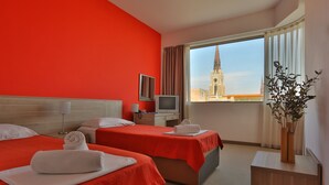 A hotel room with two beds, a TV, a window with a view of a church spire, and a plant on a side table. - Hotel Putnik (Novi Sad)