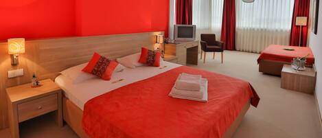 A hotel room with a large bed, a desk with a television, a chair, and a small table with a lamp.